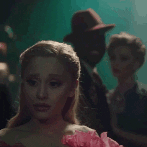 a woman in a pink dress is standing in a dark room with a man in a hat in the background .