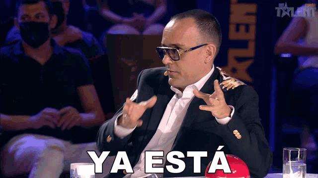 a man in a suit and sunglasses is sitting in front of a sign that says ya esta