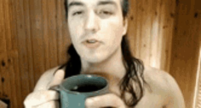 a shirtless man with long hair is drinking from a blue mug .