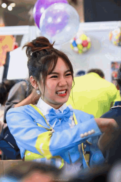 a girl wearing a blue jacket and a bow tie smiles with balloons behind her