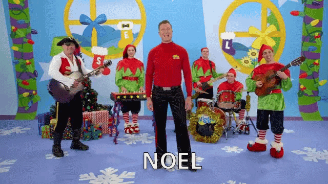 a man in a red shirt is standing in front of a band and the word noel is on the floor