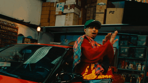 a man in a red shirt with the word fame on it stands in front of a red car in a garage
