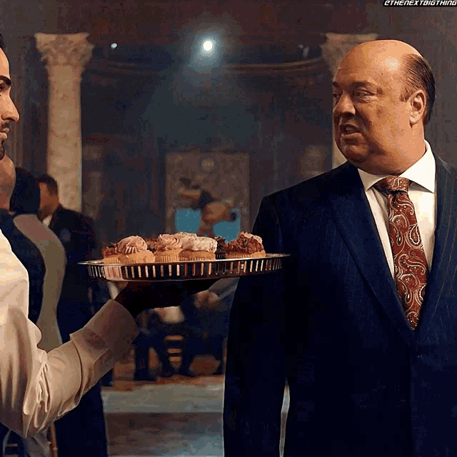 a man in a suit holds a tray of cupcakes in front of another man