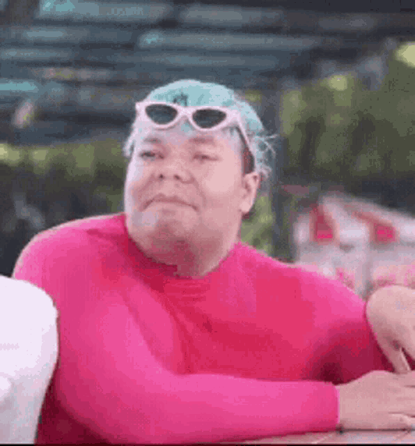 a man with blue hair and sunglasses is wearing a pink shirt and sitting at a table .