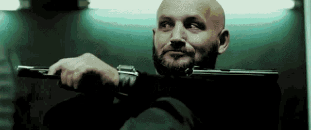 a bald man with a beard is holding a gun over his shoulder