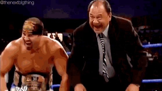 a man in a suit and tie is pointing at a wrestler in a ring .
