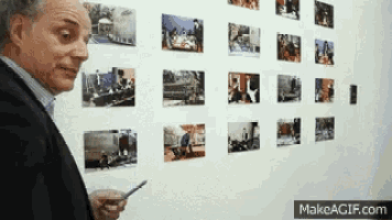 a man in a suit is looking at pictures on a wall with makeagif.com at the bottom of the screen