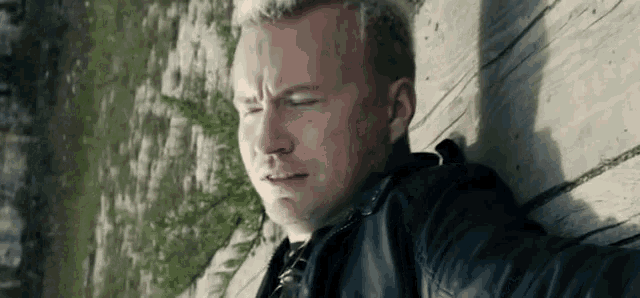 a man is leaning against a wooden wall with his eyes closed and a serious look on his face .