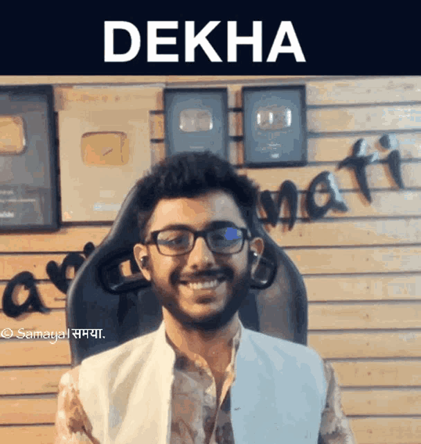 a man wearing glasses and a white vest is smiling in front of a wall with the word dekha on it