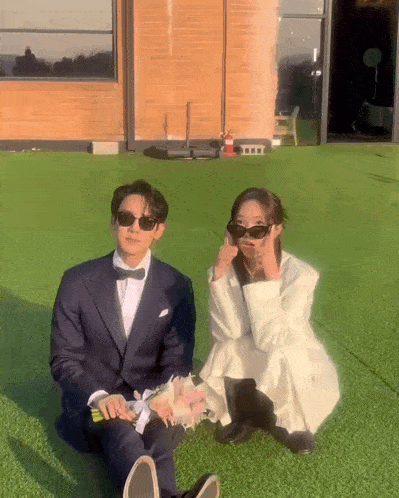 a man in a tuxedo and a woman in a white dress are sitting on the grass