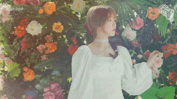 a woman in a white dress is standing in front of a wall of flowers with the word kiwi pop on the bottom