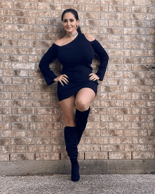 a woman standing in front of a brick wall wearing a black dress and black boots