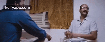 two men are sitting in a room talking to each other in front of an air conditioner .