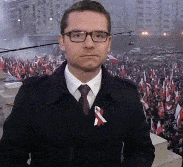 a man wearing glasses stands in front of a crowd with flags and a red and white ribbon around his neck