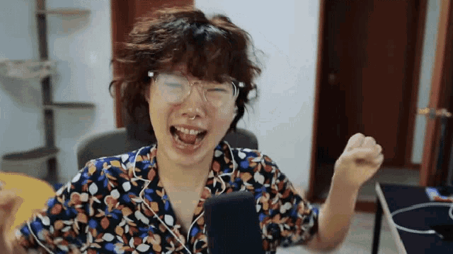 a woman wearing glasses and a floral shirt laughs with her fist in the air