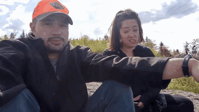 a man wearing an orange hat with a bear on it sits next to a woman wearing a watch