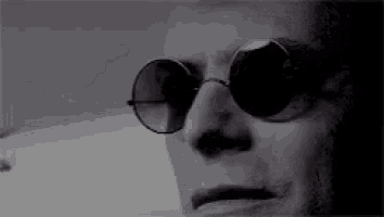 a man wearing round sunglasses is looking at the camera in a black and white photo .