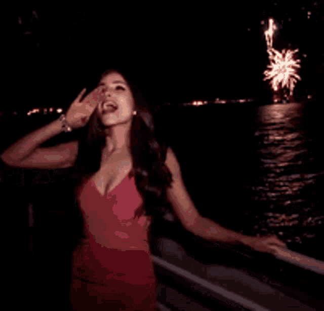 a woman in a red dress stands in front of a fireworks display over a body of water