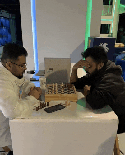 two men are playing a game of chess at a table that has a sign that says hills bank