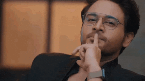 a man wearing glasses and a watch is holding his finger to his lips .