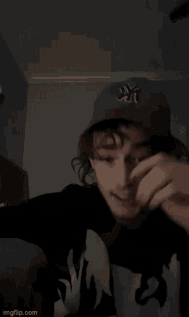 a young man wearing a ny hat and a black hoodie is covering his face with his hand .