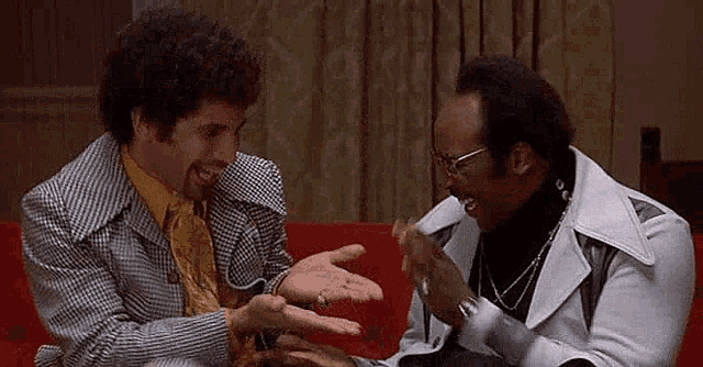 two men are sitting on a red couch laughing .