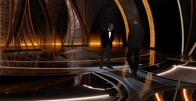 two men in tuxedos are dancing on a stage in a dark room