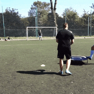 a man in a black shirt stands on a soccer field watching another man kick a ball