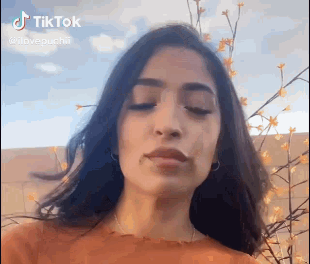 a woman with long dark hair is standing in front of a tree with flowers and making a funny face .