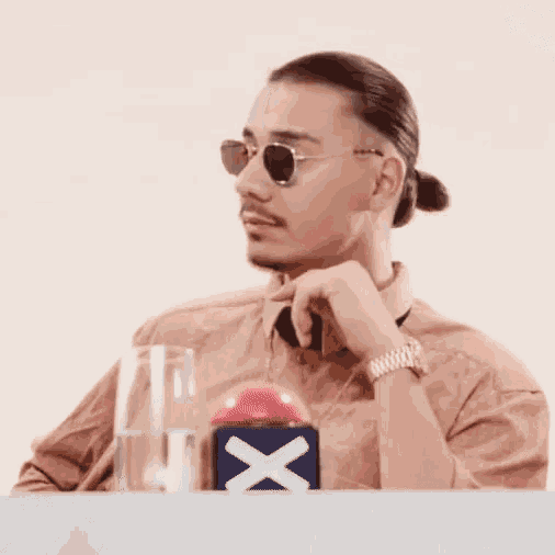a man wearing sunglasses and a ponytail sits at a table with a glass of water