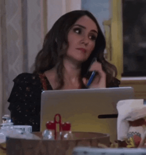 a woman is sitting at a table talking on a cell phone while using a laptop .