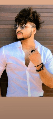 a young man wearing sunglasses and a white shirt stands in front of a wooden wall .
