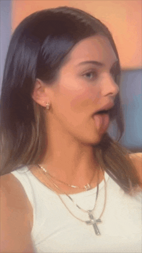 a woman wearing a white tank top and a cross necklace sticks her tongue out