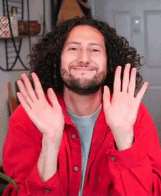 a man with curly hair and a beard is wearing a red shirt and waving his hands .