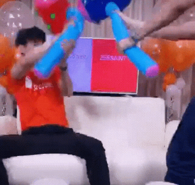 a man in an orange shirt is sitting on a couch holding balloons in front of a television screen .