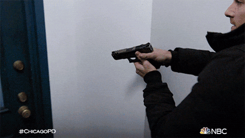 a man is holding a gun in front of a white wall with nbc written on the bottom right