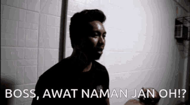 a man in a black shirt is standing in front of a white wall with the words `` boss , awat nama jan oh ? ''
