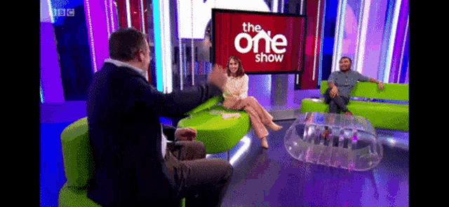 a man and a woman sit on a green couch in front of a screen that says the one show