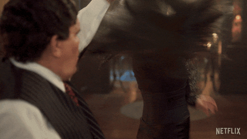 a woman in a black dress is dancing with a man in a suit and tie in front of a netflix logo