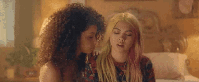 two women are sitting next to each other on a bed . one of the women has pink hair .