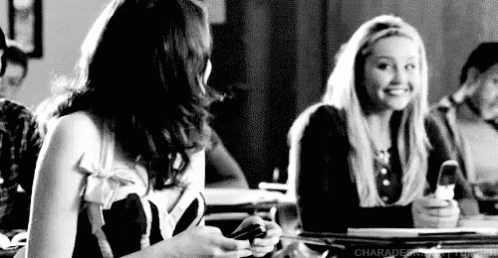 a black and white photo of two girls sitting at a table looking at their phones .
