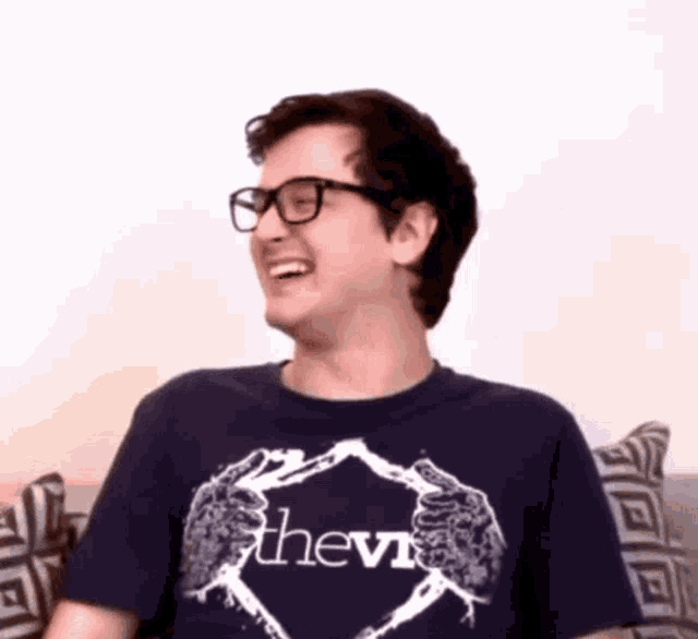 a man wearing glasses and a blue shirt with the word thevi on it is laughing while sitting on a couch .