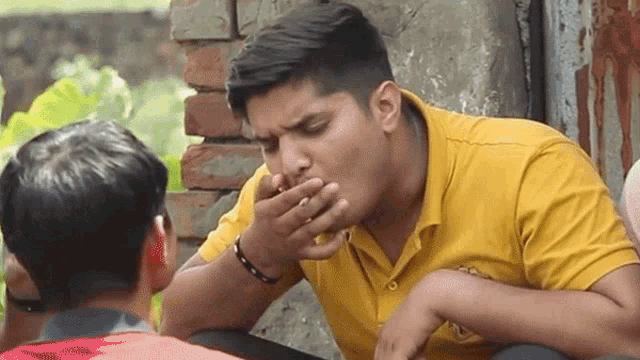 a man in a yellow shirt is covering his mouth with his hand while sitting in front of a brick wall .