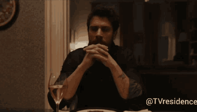 a man with a beard sits at a table with a glass of wine in front of a tv residence logo