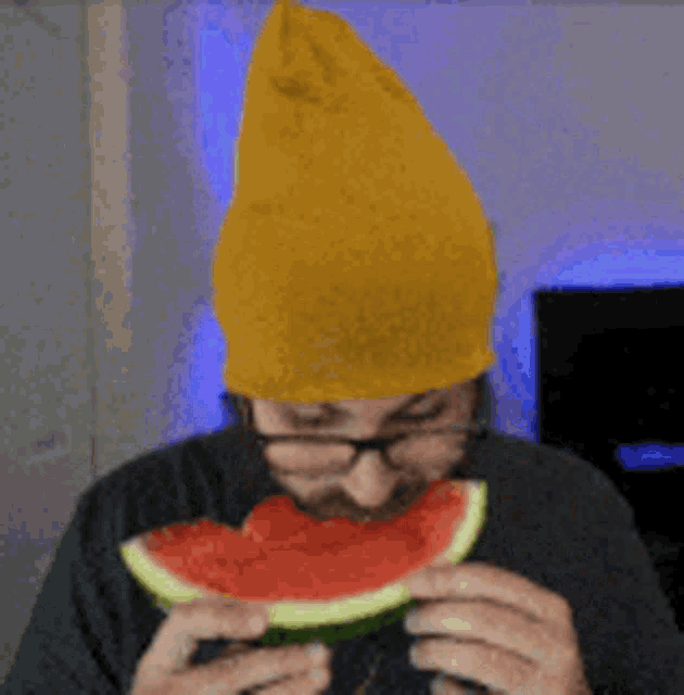 a man wearing glasses and a yellow beanie is eating a slice of watermelon