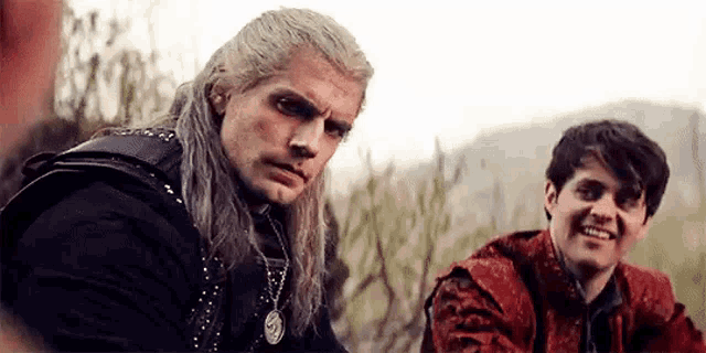 a man with long hair is sitting next to another man in a field .