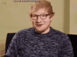 a man wearing glasses and a blue sweater is sitting in a chair and smiling .