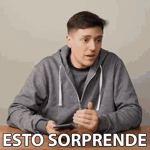 a man in a grey hoodie is sitting at a table holding a cell phone with the words esto sorprende behind him