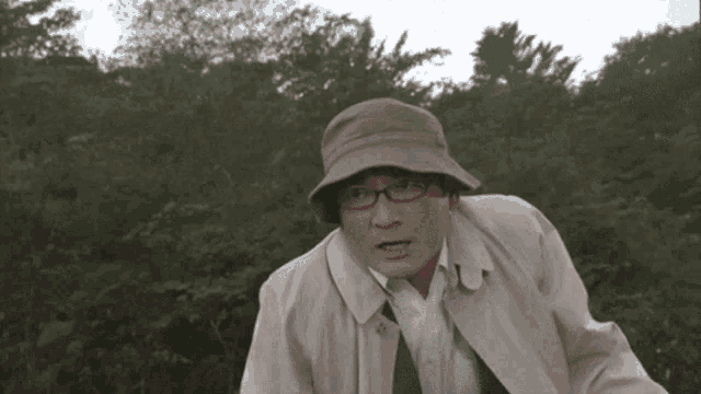 a man wearing glasses and a hat stands in front of a forest