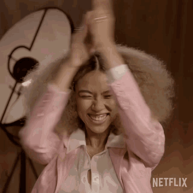 a woman wearing a pink jacket and a white shirt is clapping her hands in front of a netflix logo